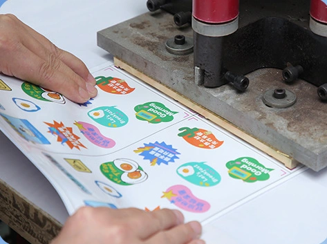 Hands positioning a printed sticker sheet beneath a production press