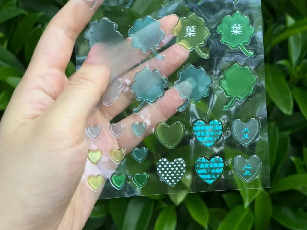 A clear puffy sticker sheet held against foliage, with fingers and background visible through clear areas.
